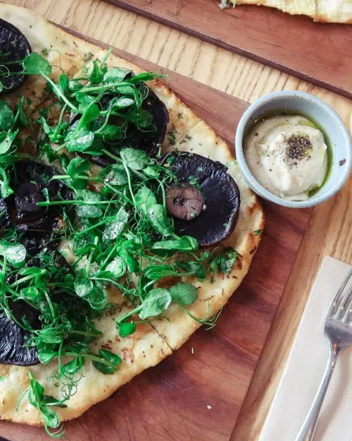 A mushroom and rocket pizza on a wooden board with a bowl of houmous dip