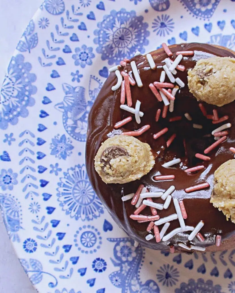 Overhead photo of a chocolate glazed doughnuts with pink and white sprinkles and mini balls of cookie dough on a blue and white patterned plate