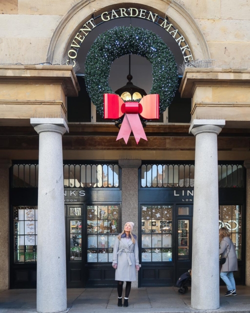 Woman in long grey coat and wooly hat stood under Covent Garden Christmas sign and wreath