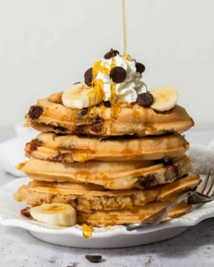 A stack of golden vegan chocolate chip waffles topped with dairy free cream, chocolate chips and syrup