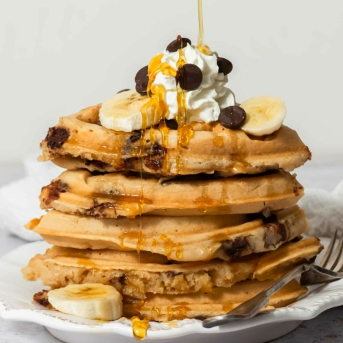A stack of golden vegan chocolate chip waffles topped with dairy free cream, chocolate chips and syrup