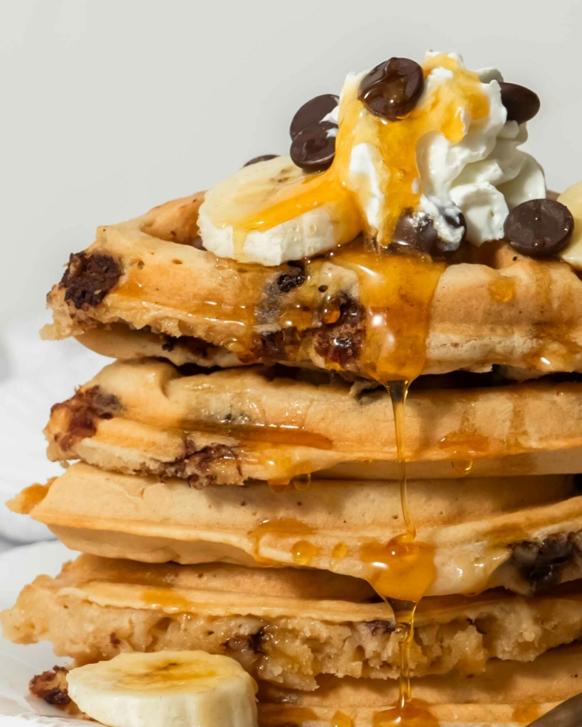 A stack of golden vegan chocolate chip waffles topped with dairy free cream, chocolate chips and syrup