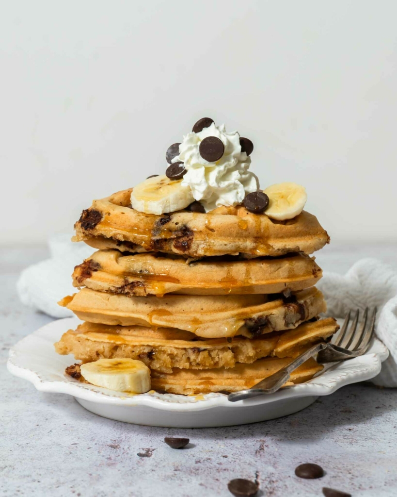 A stack of golden vegan chocolate chip waffles topped with dairy free cream, chocolate chips and syrup