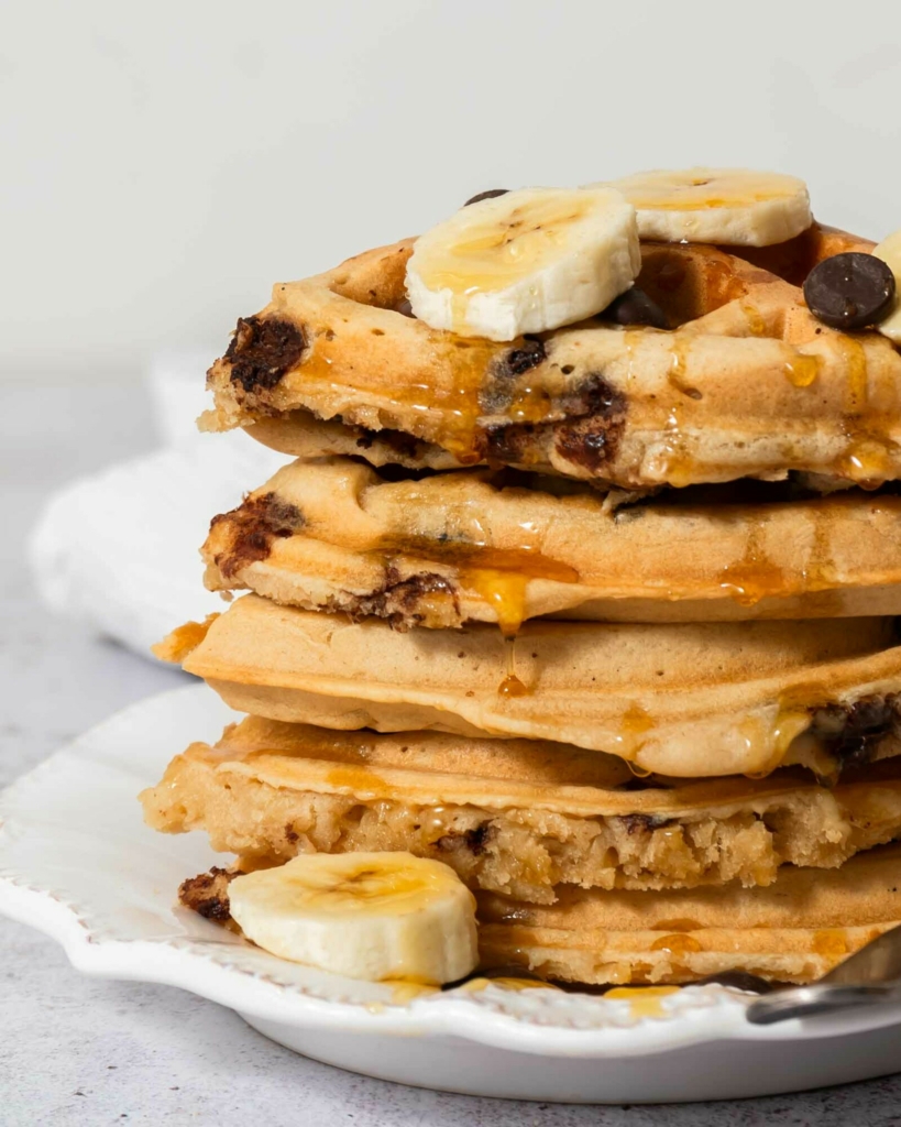 A stack of vegan waffles with banana slices, chocolate chips and syrup