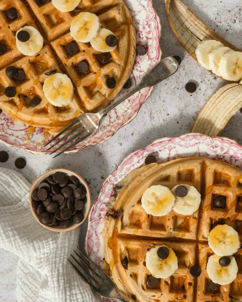 An overhead photograph of two large vegan waffles topped with banana slices, syrup and chocolate chips