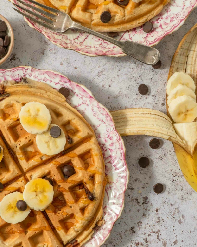 An overhead photograph of two large vegan waffles topped with banana slices, syrup and chocolate chips