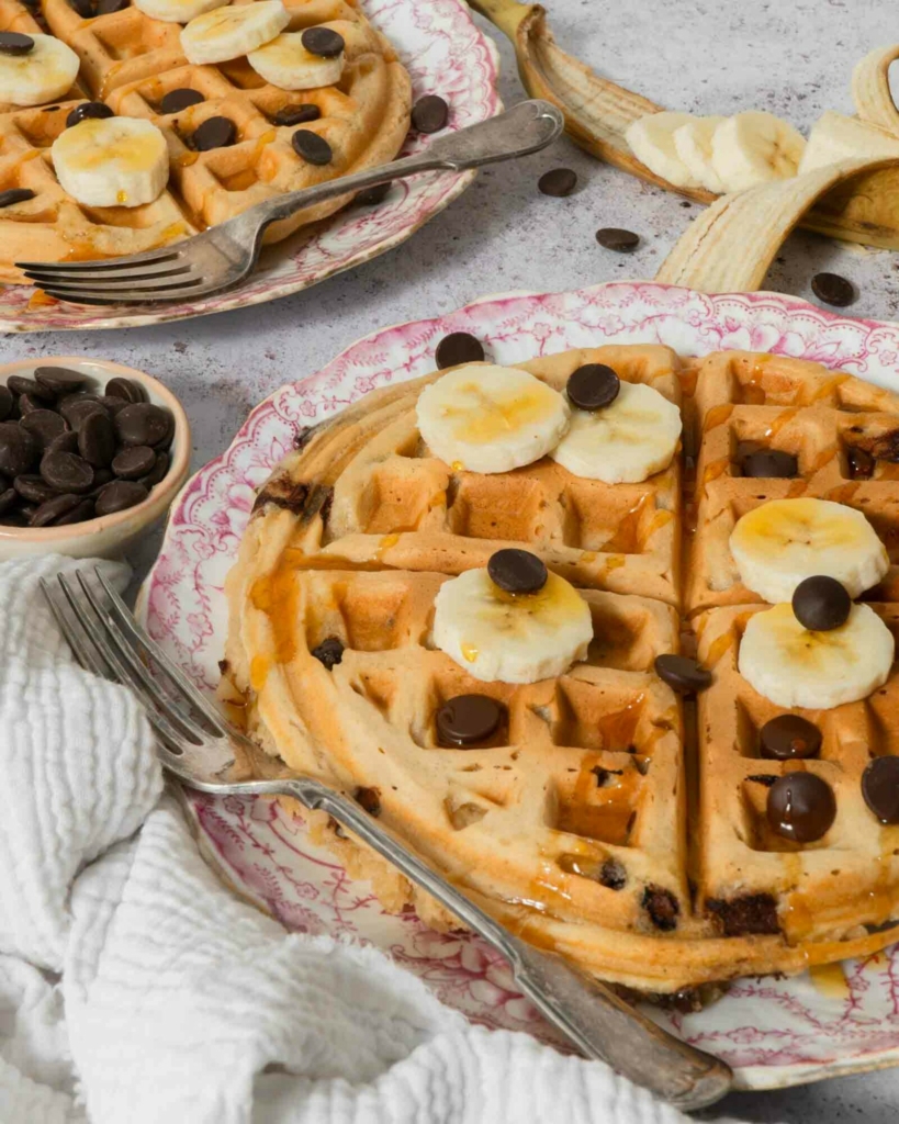 An overhead photograph of two large vegan waffles topped with banana slices, syrup and chocolate chips