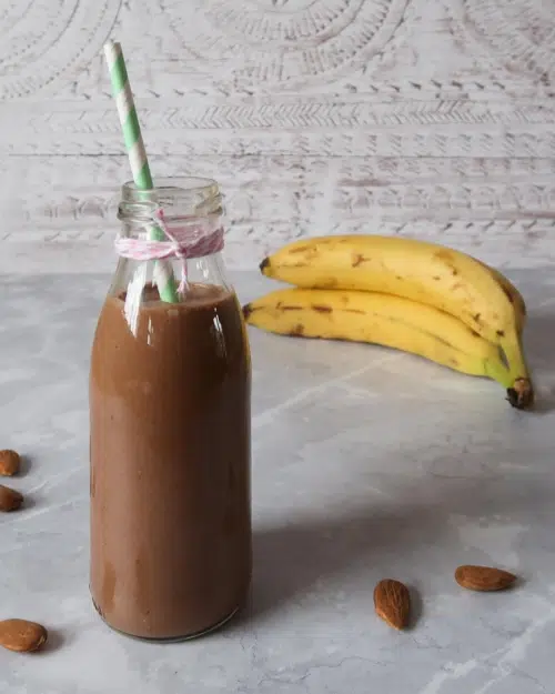 Miniature milk bottle of a vegan healthy banana chocolate milkshake with a stripy straw and fresh bananas in the background