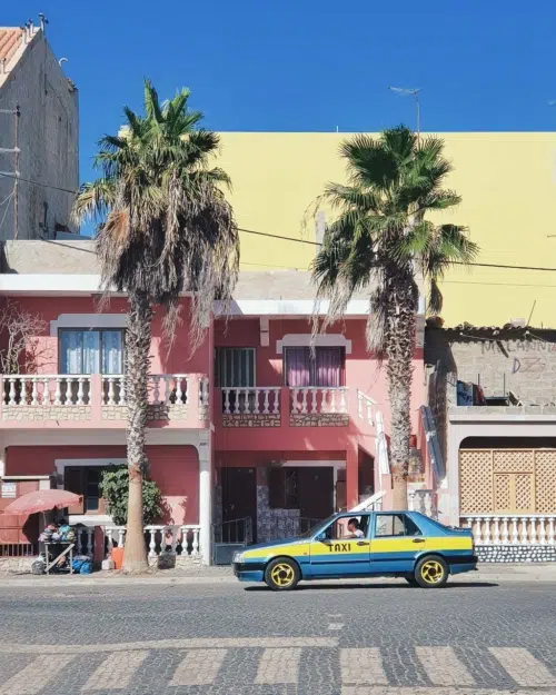 Blue and yellow taxi on the street next to a pink and yellow building in Cape Verde