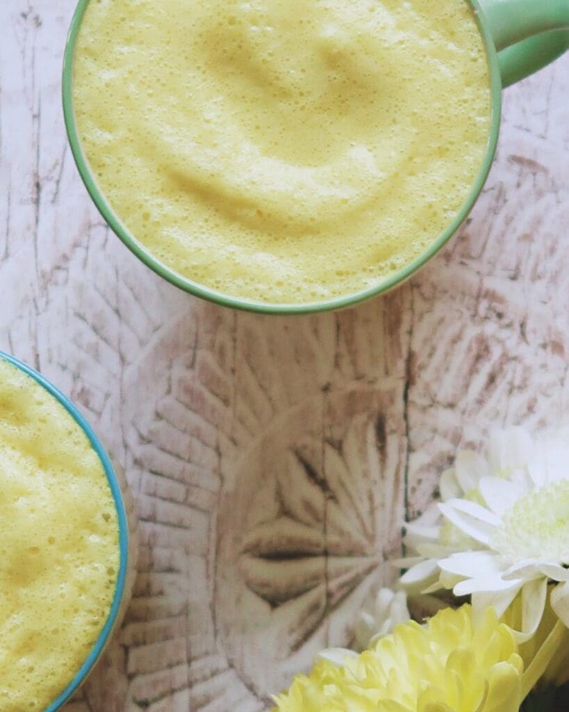 Flat lay of two cups of turmeric latte, with pastel yellow milk foam and a bunch of yellow and white flowers beside