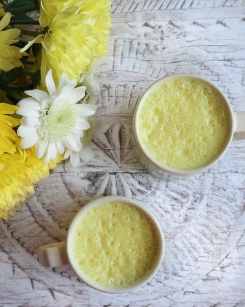 Flat lay of two cups of turmeric latte, with pastel yellow milk foam and a bunch of yellow and white flowers beside