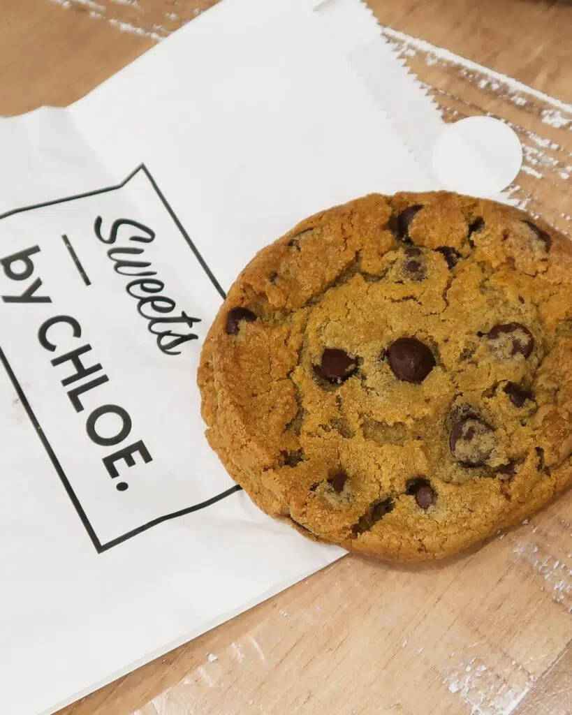 A vegan gluten free chocolate chip cookie sat on a 'By Chloe' white paper bag
