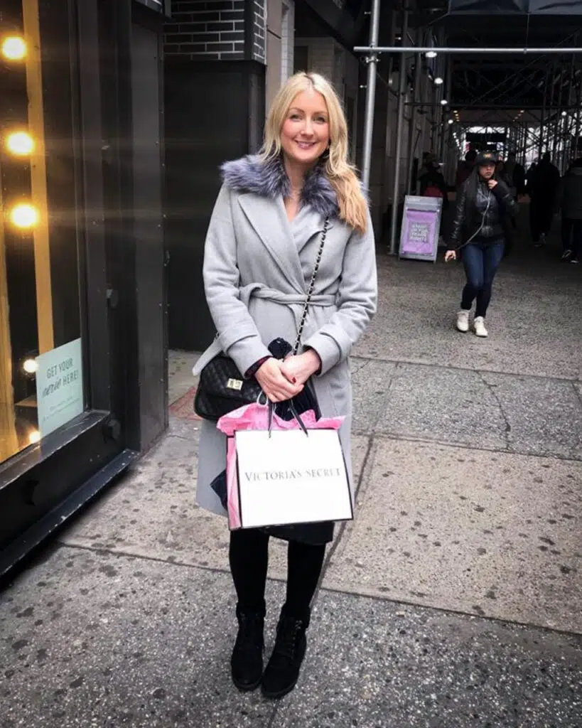 A blonde woman in a long grey coat with faux fur trim, standing outside on the street in New York City holding shopping bags and smiling