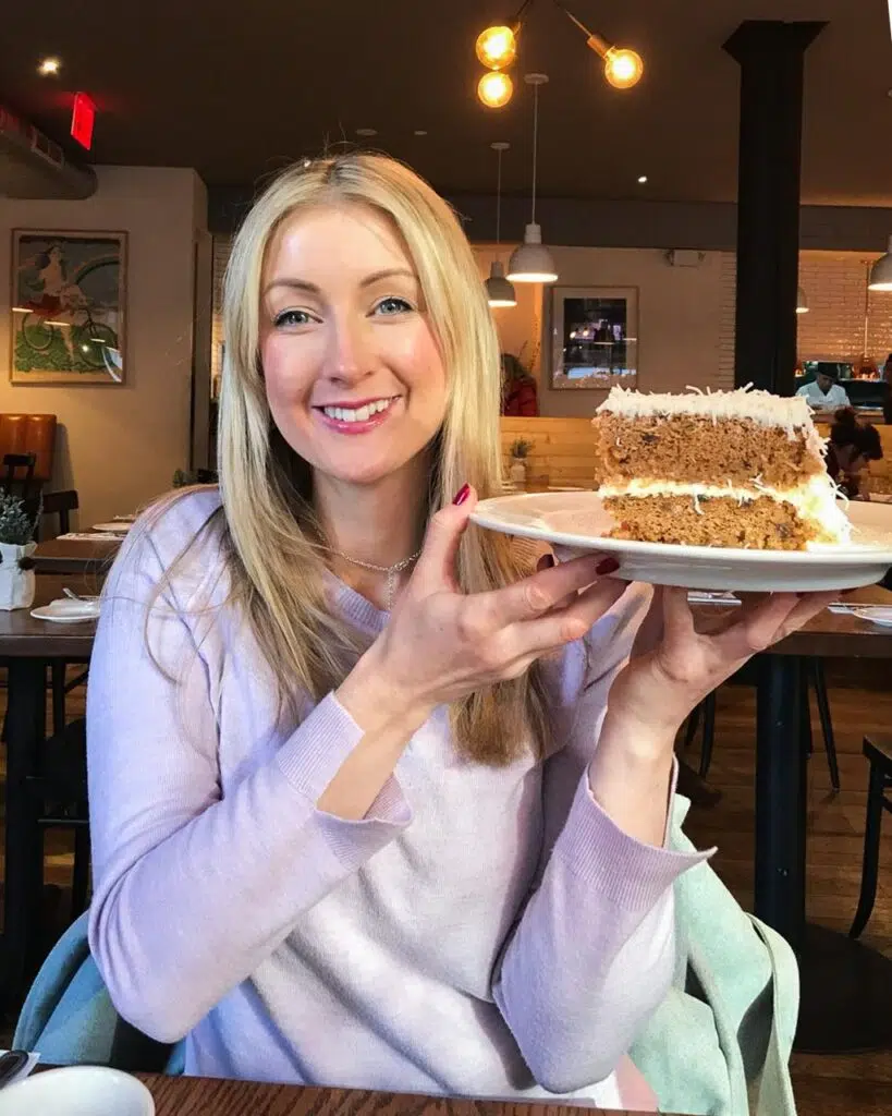 A blonde woman in a lilac jumper smiling and holding a large slice of vegan carrot cake in Spring Street Naturals restaurant in New York City