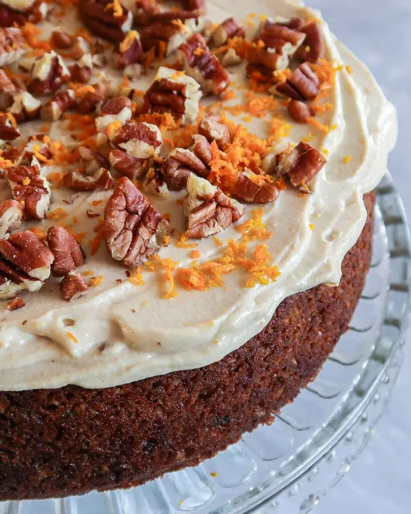 A rich carrot cake standing on an glass cake stand, topped with tofu icing, grated orange zest and toasted pecans