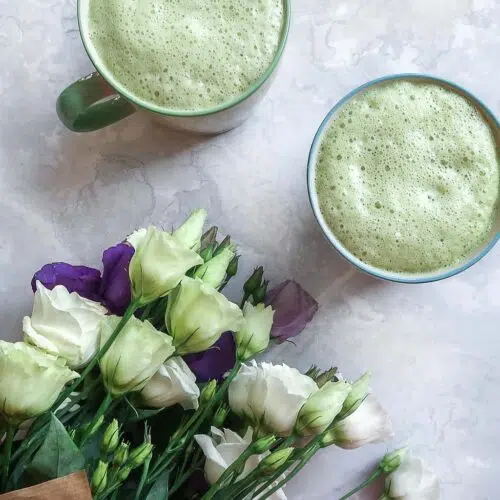 Two cups of frothy green matcha latte next to a bunch of white and purple flowers