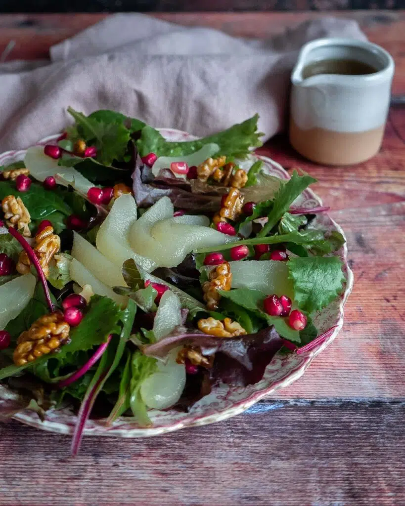 A poached pear, pomegranate and candied walnut salad sat on a pretty pink patterned floral plate with a small jug of salad dressing and a napkin behind