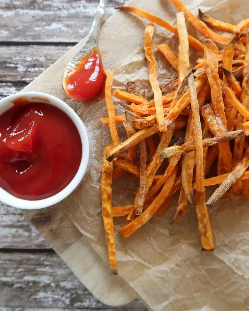 Skinny sweet potato fries on a paper and rustic wood background with a round dish of ketchup
