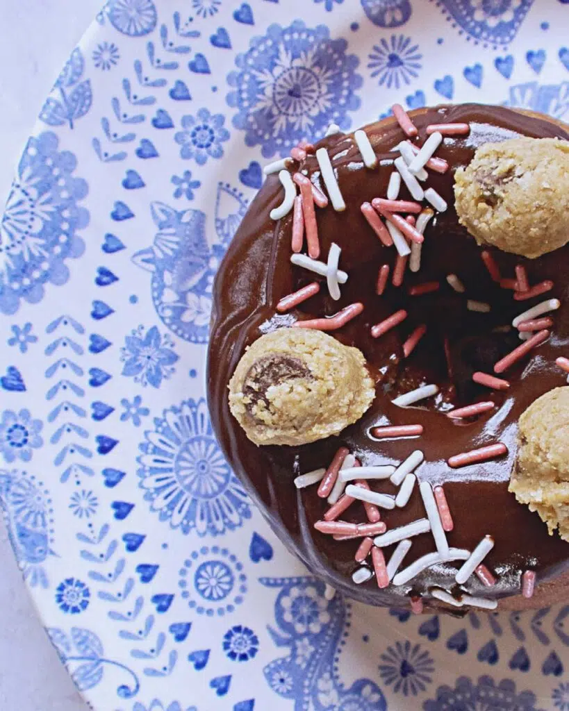 A chocolate coated doughnut on a blue patterned plate with balls of vegan cookie dough on top