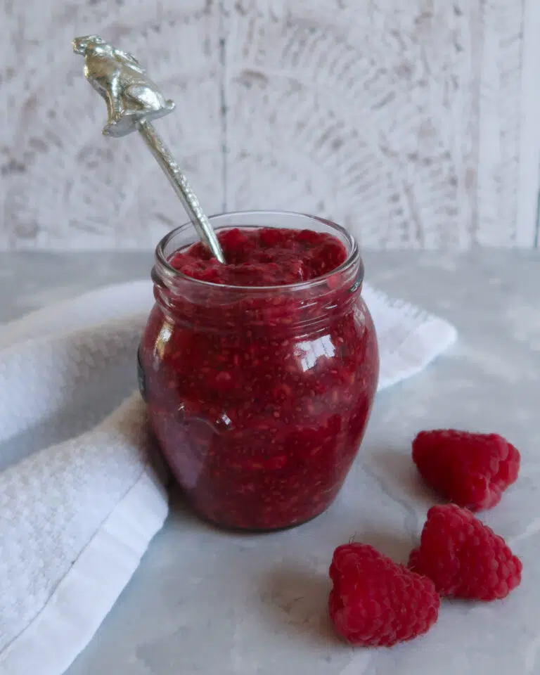 A small jar of red raspberry chia seed jam with fresh raspberries decorating the scene
