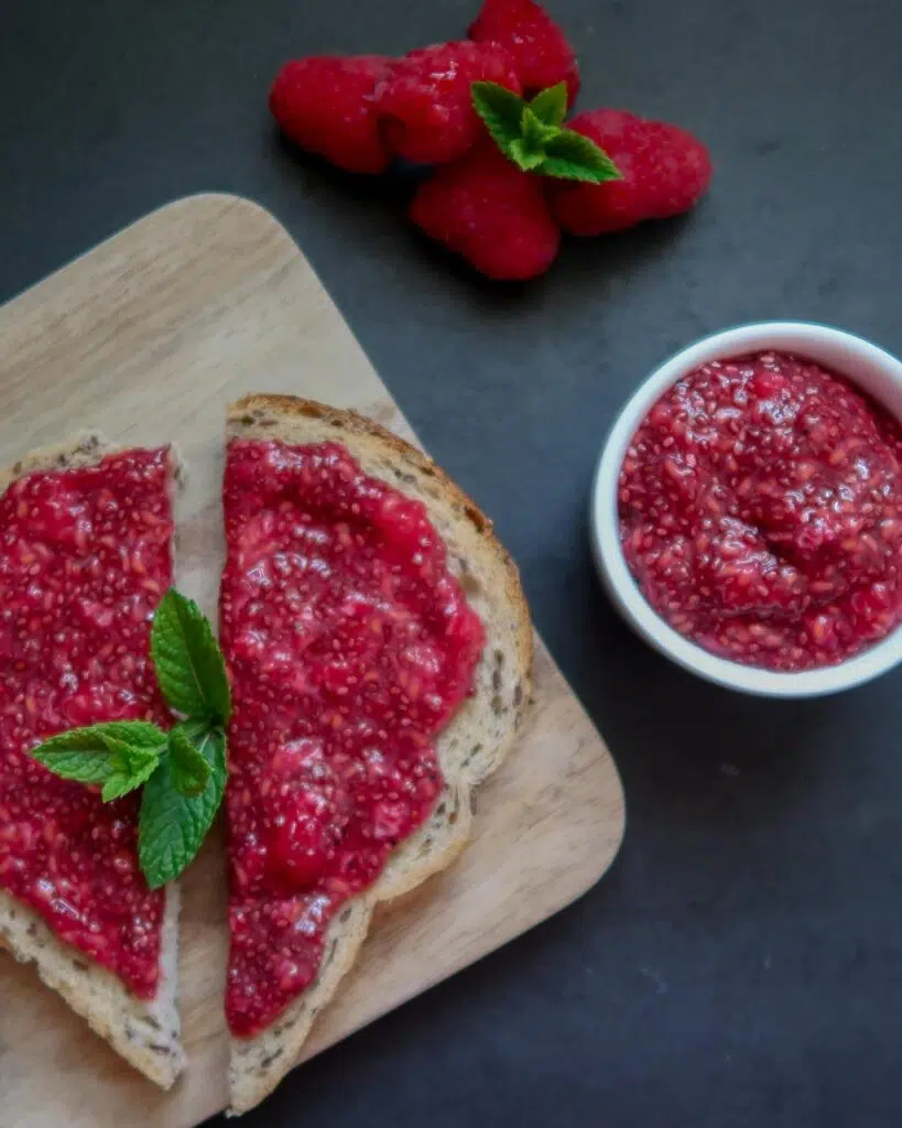 A slice of wholemeal toast with red raspberry chia seed jam spread generously on top and garnished with a fresh mint leaf.