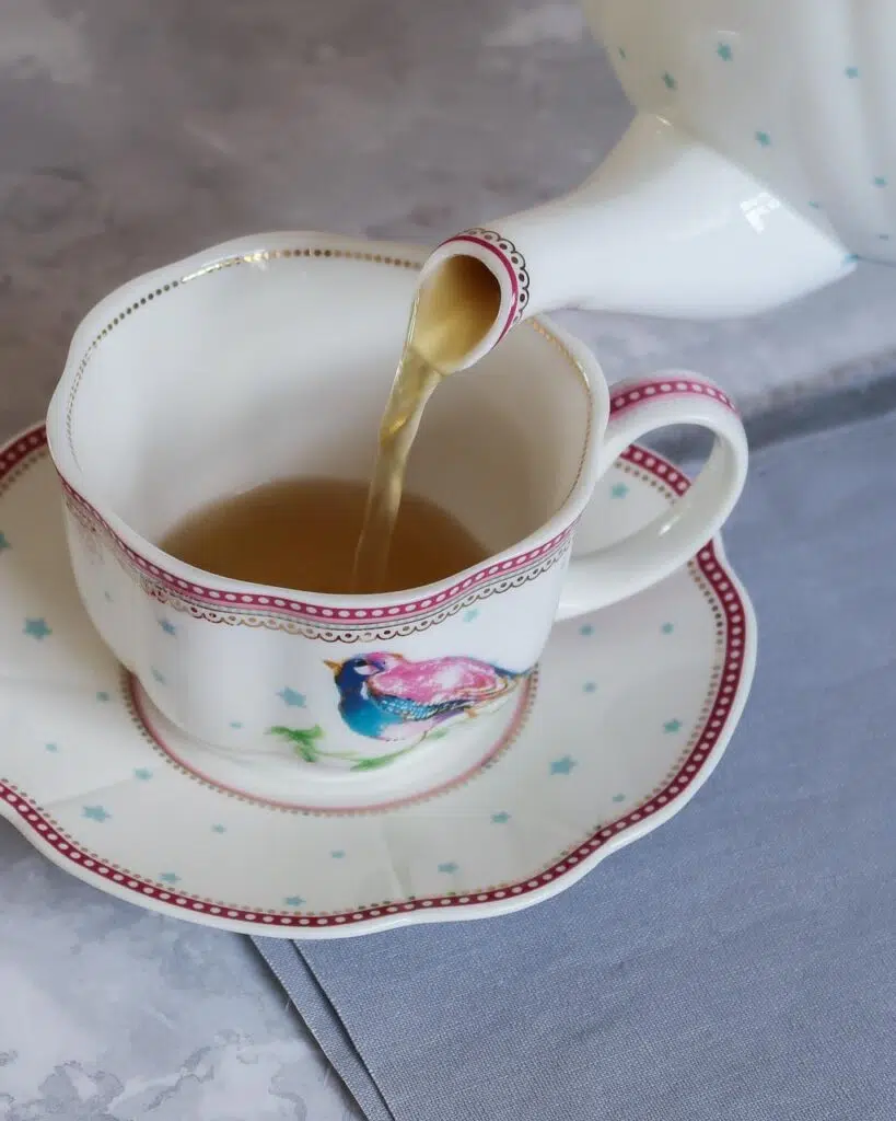 A tea pot pouring green tea into a white tea cup sat on a saucer, decorated with a pink and blue bird