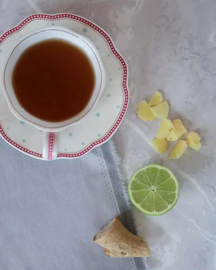 A teacup on a saucer holding a cup of tea, with half a lime and fresh ginger slices near by