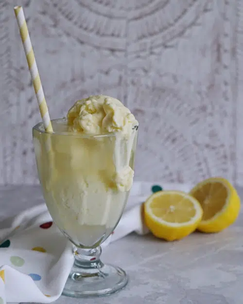 A glass of lemonade with coconut ice cream scooped on top, melting down the sides of the glass, with fresh lemons in the background and a yellow and white striped paper straw