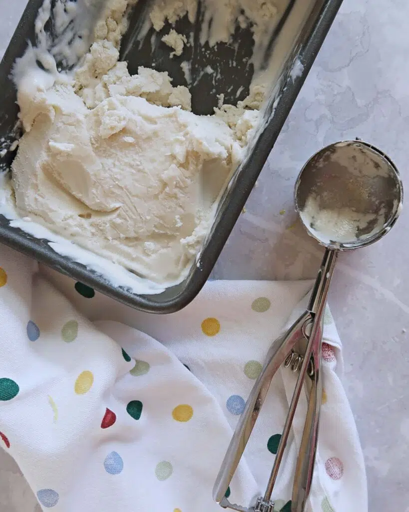 A dish of homemade coconut ice cream sat on a polka dot tea towel and a silver metal ice cream scoop