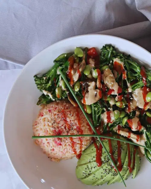 A bowl of vegan stir fry with fresh green tenderstem broccoli, edamame and red peppers served with a creamy peanut butter sauce, red chilli sauce, pink speckled rice and avocado