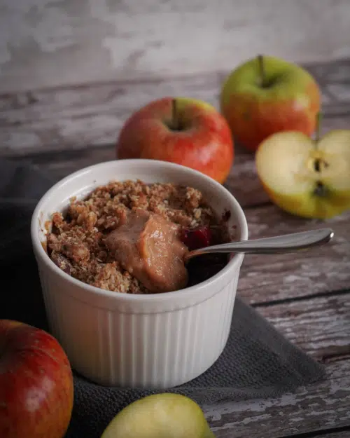 Mini apple crumble pot with a caramel centre on a wooden backdrop with fresh green and red apples surrounding it