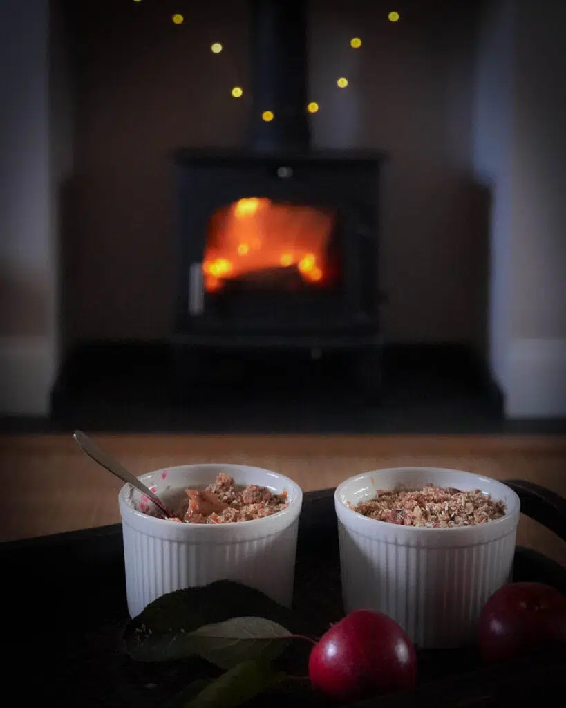 Mini apple crumble pots on a tray in front of a roaring fire with fairy lights adorning the fireplace