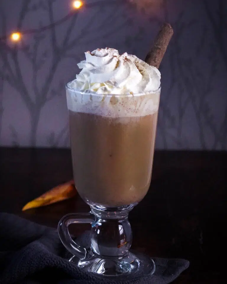A tall glass mug filled with vegan pumpkin spice latte and topped with vegan spray whipped cream and a cinnamon stick