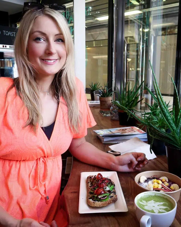 Blonde woman in an orange dress sat at a counter in a cafe with an avocado toast, matcha latte and acai bowl
