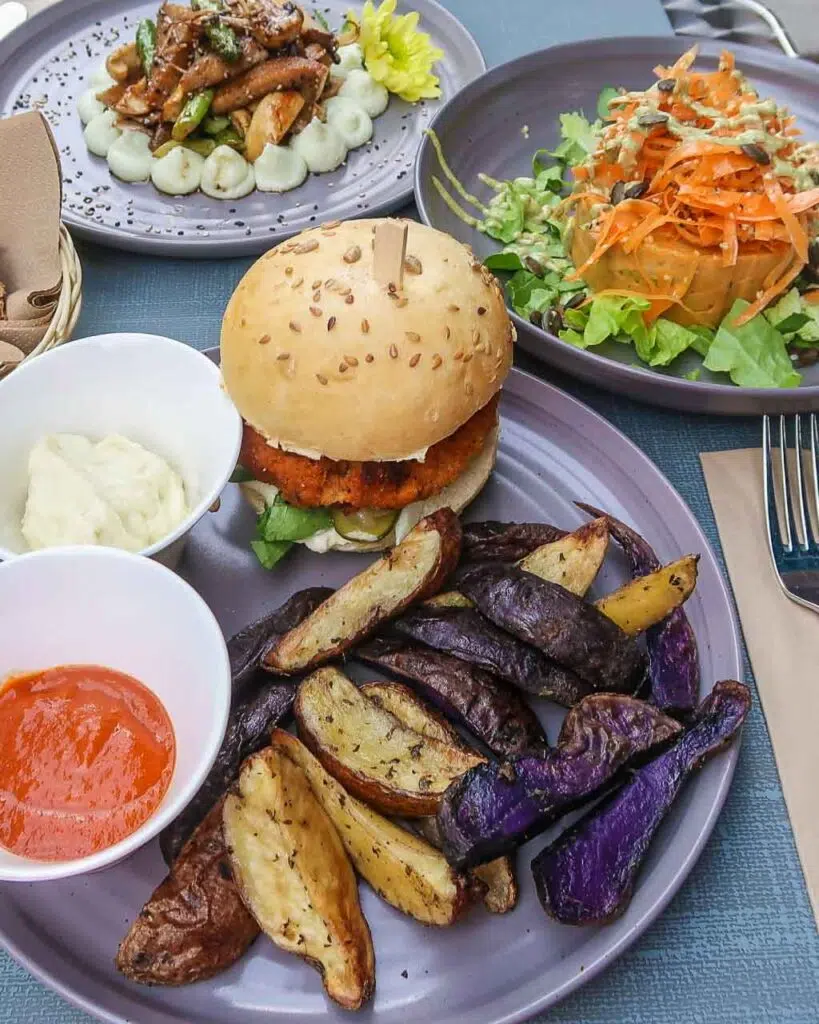 Three plates of vegan food including a burger and bun with purple and white potato wedges, a carrot salad and a stir fry with sides