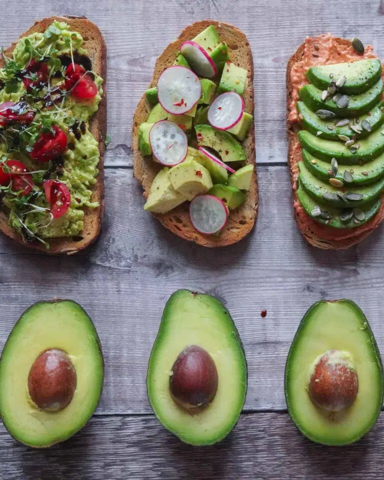 A flat lay of three slices of avocado toast with different garnishes and toppings and three open ripe avocado halves with their stones showing