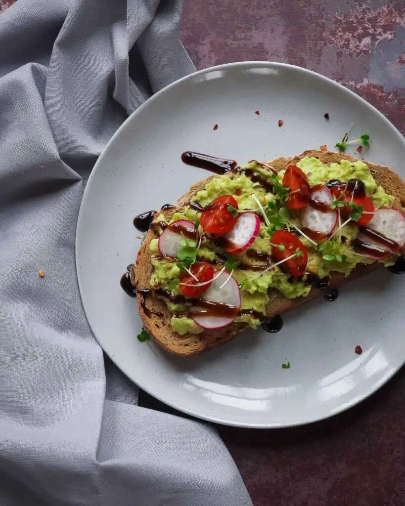 A slices of sourdough toast with mashed avocado, cherry tomatoes, radish slices, sprouts and balsamic glaze drizzled on top