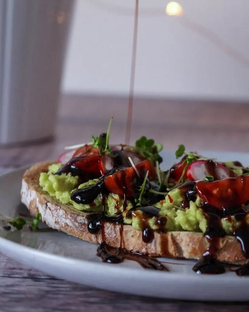 A slices of sourdough toast with mashed avocado, cherry tomatoes, radish slices, sprouts and balsamic glaze being drizzled on top