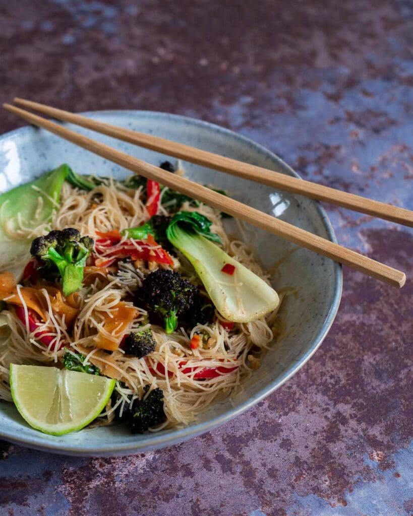 A bowl of vegan stir fry with fresh vegetables and noodles with chop sticks on top