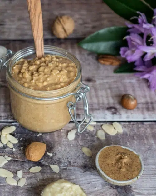A glass clip top jar full of peanut butter with various nuts surrounding it, a smaller dish of almond butter and some purple flowers
