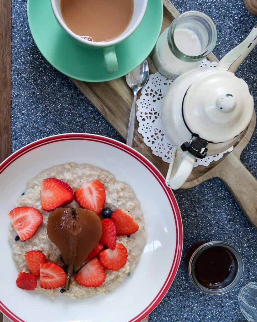 An overhead shot of a bowl of porridge with a poached pear and fresh strawberries on top, with a teapot and cup of tea on the side