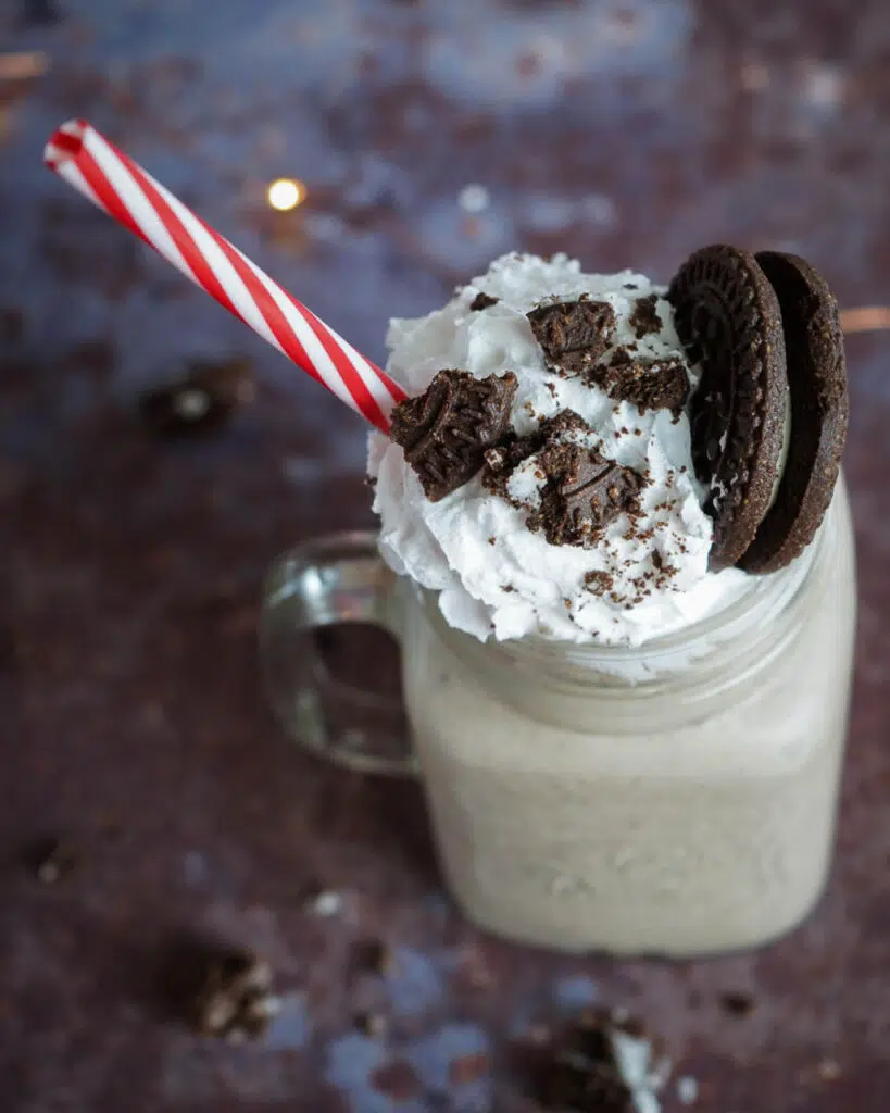 A glass mug with handle holding a cookies and cream milkshake, topped with whipped cream, an Oreo cookie, crumbled Oreos and with a red and white stripe reusable straw