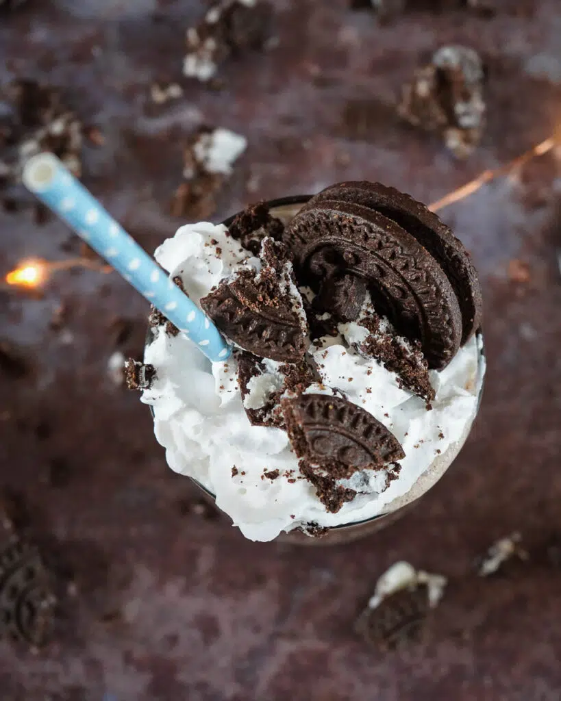 A top down shot of a vegan milkshake topped with whipped cream, an Oreo, crumbled cookies and a blue and white paper straw on a dark background with fairy lights and crumbled cookies