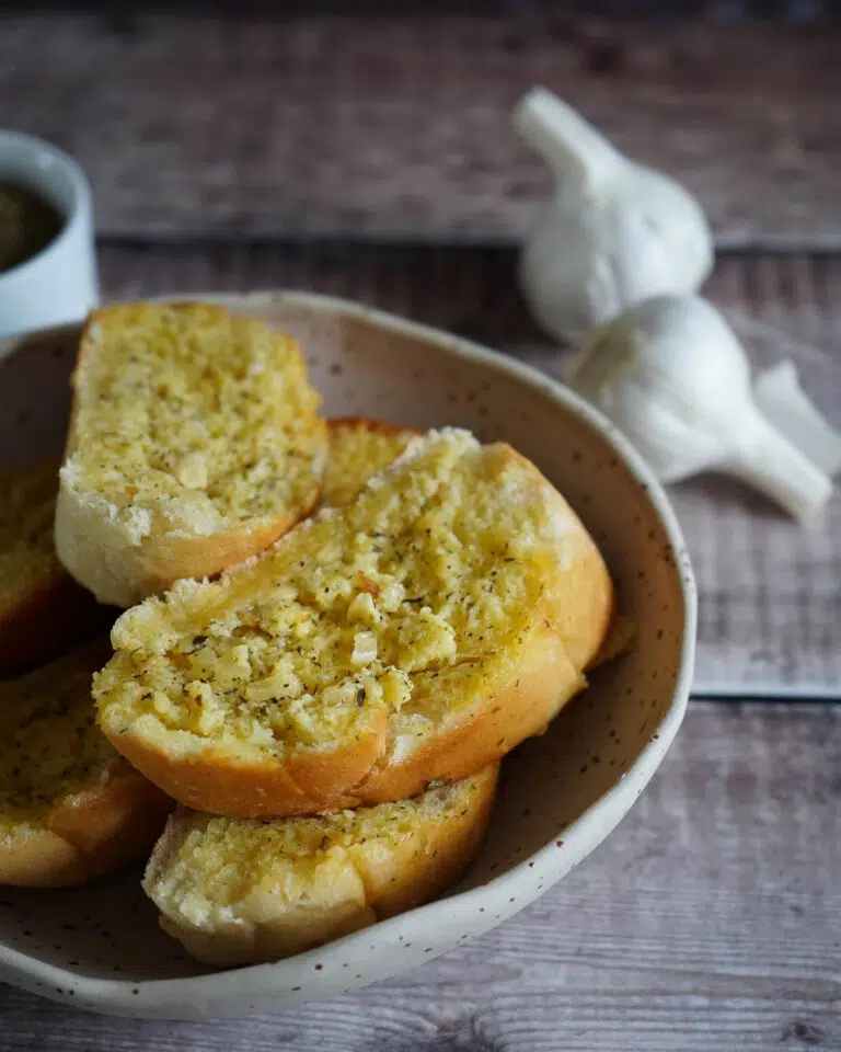 A bowl of garlic bread slices with two whole garlic bulbs out of focus in the background