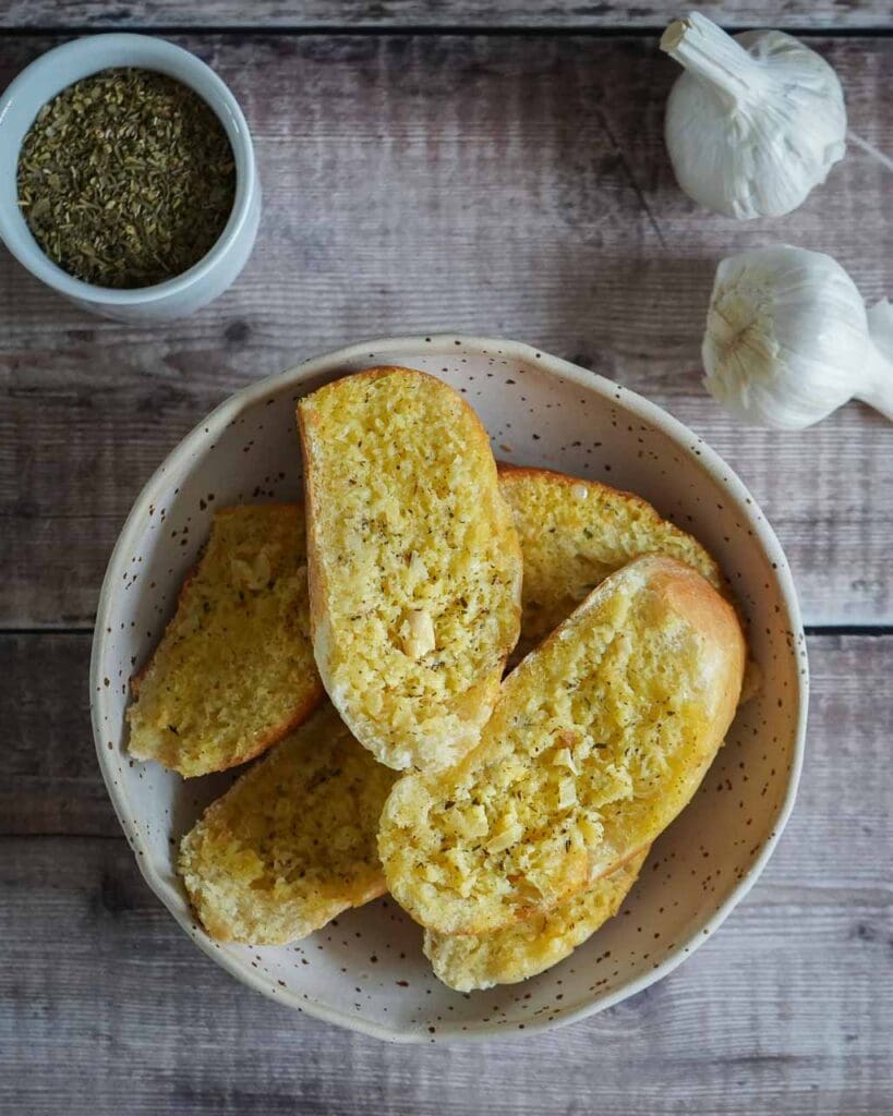 A bowl of garlic bread slices with two whole garlic bulbs set behind with a small dish of dried herbs