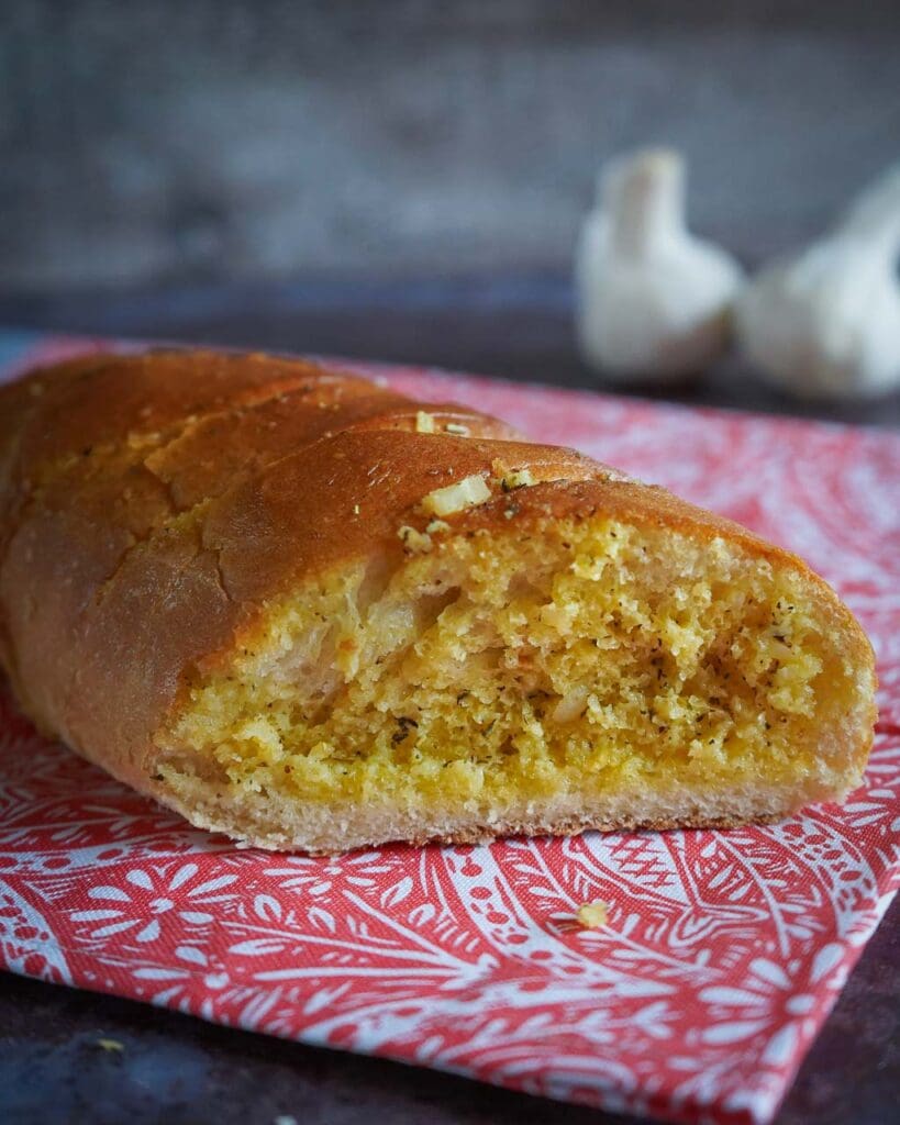 A golden garlic baguette fresh from the oven set on a red and white patterned tea towel, with two small garlic bulbs out of focus in the background