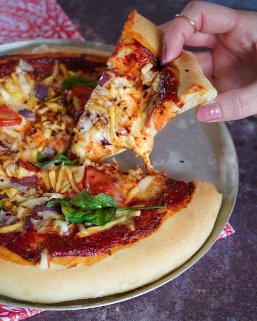 A colourful vegan pizza with one slice being lifted out and a cheese string forming