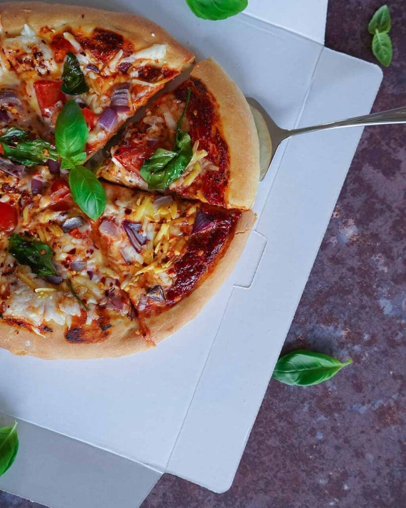 A colourful vegan pizza sat on a cardboard pizza box with fresh basil leaves sprinkled around