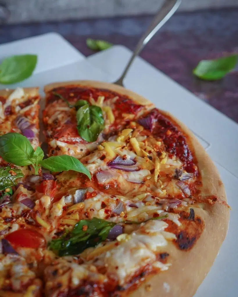 A colourful vegan pizza sat on a cardboard pizza box with fresh basil leaves sprinkled around