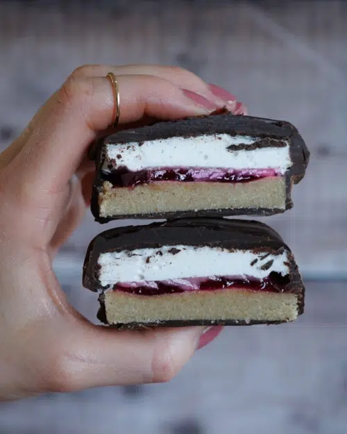 A hand holding two halves of a vegan wagon wheel one on top of the other, revelling the layers of biscuit, jam and marshmallow